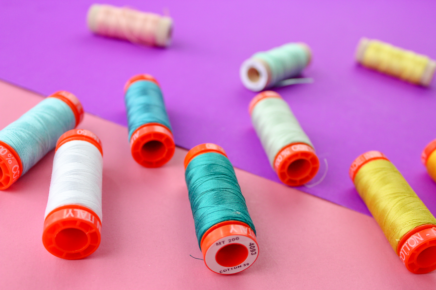Pictured: varying colors of thread on spools. On a pink and purple background.